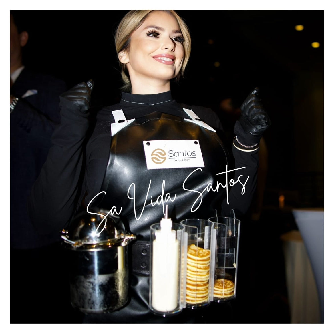 Caviar Girl from Santos Gourmet smiling while presenting caviar, blinis, and sauce at a social gathering, adding elegance and flair.